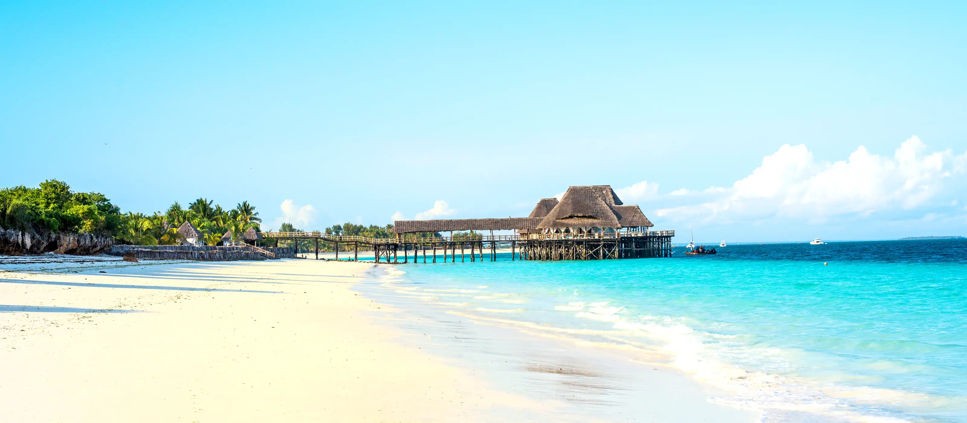 A wide empty beach at low tide with a thatched jetty in the distance