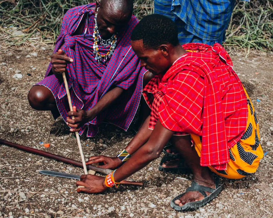 Two Maasai men crouched on the ground spinning a stick between their palms to make fire