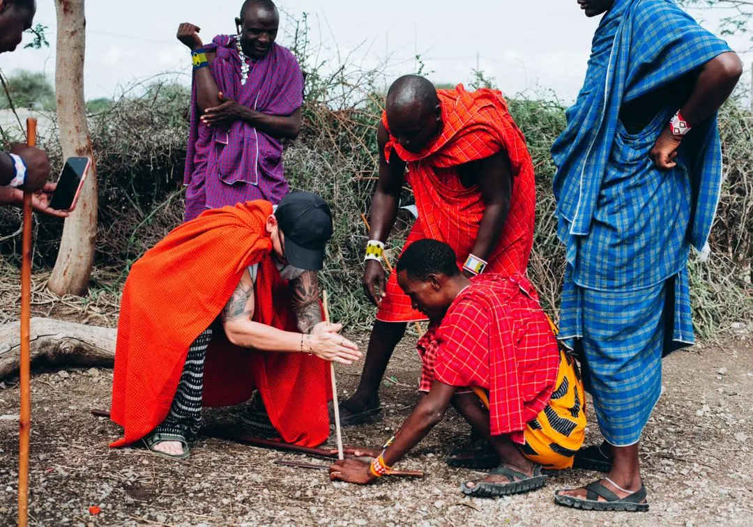 A guest in a red shuka trying the fire stick, Maasai men guiding and watching
