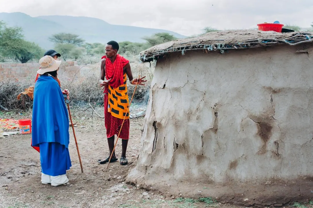 A guest in a blue shuka listening to a Maasai man in red outside a mud-walled hut, hills behind