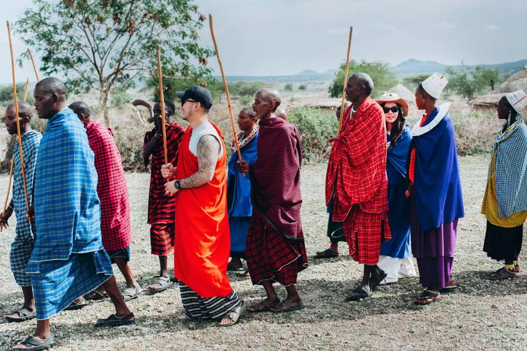 Maasai men and women in shukas walking in line with their sticks, two guests among them, the plain and hills beyond