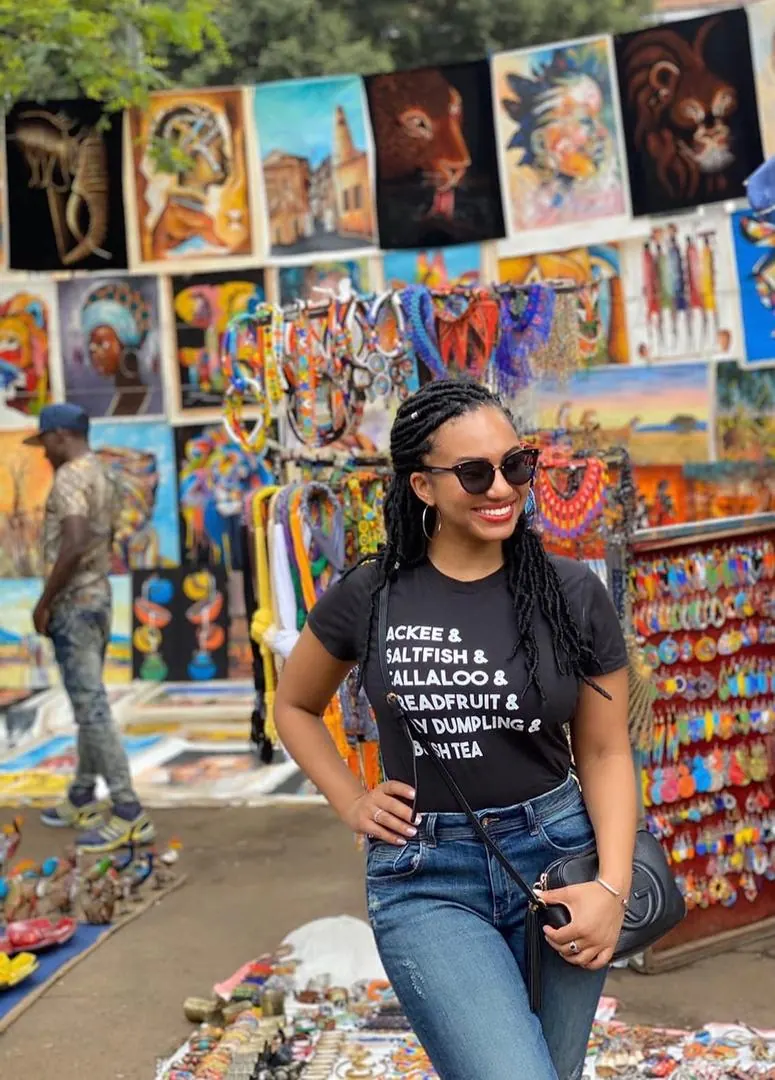 A visitor smiling among the paintings, beadwork and carvings hung along a market stall