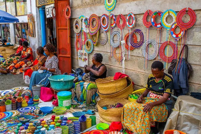 Women selling beadwork at a street market, beaded collars hanging on the wall behind them