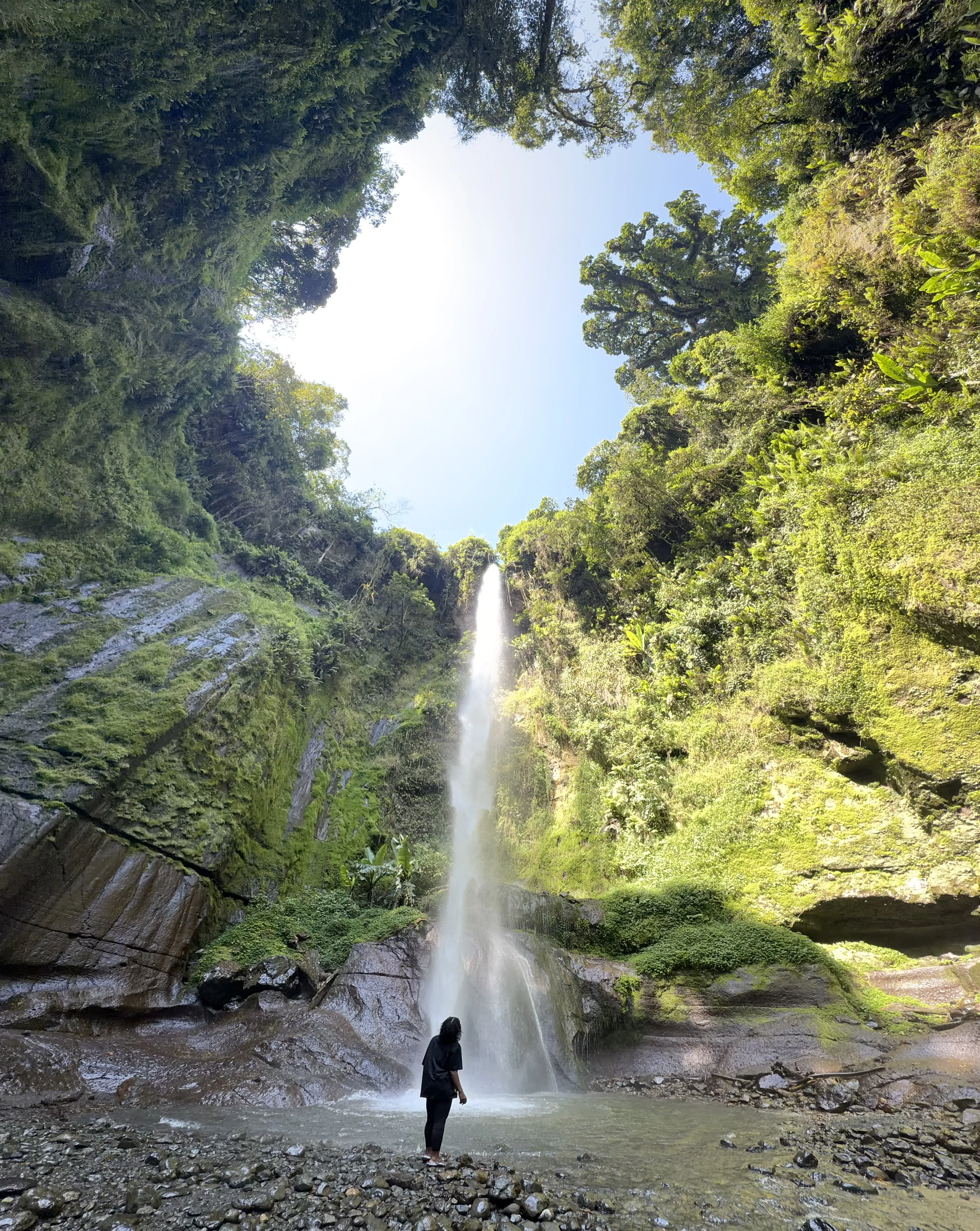 A visitor standing at the foot of Napuru waterfall, small beneath the forested gorge walls