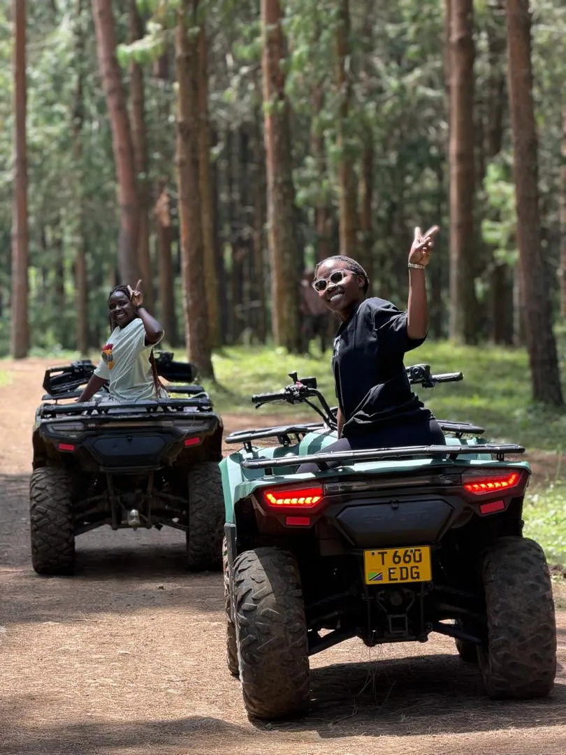 Two riders on quad bikes waving from a track through a pine forest