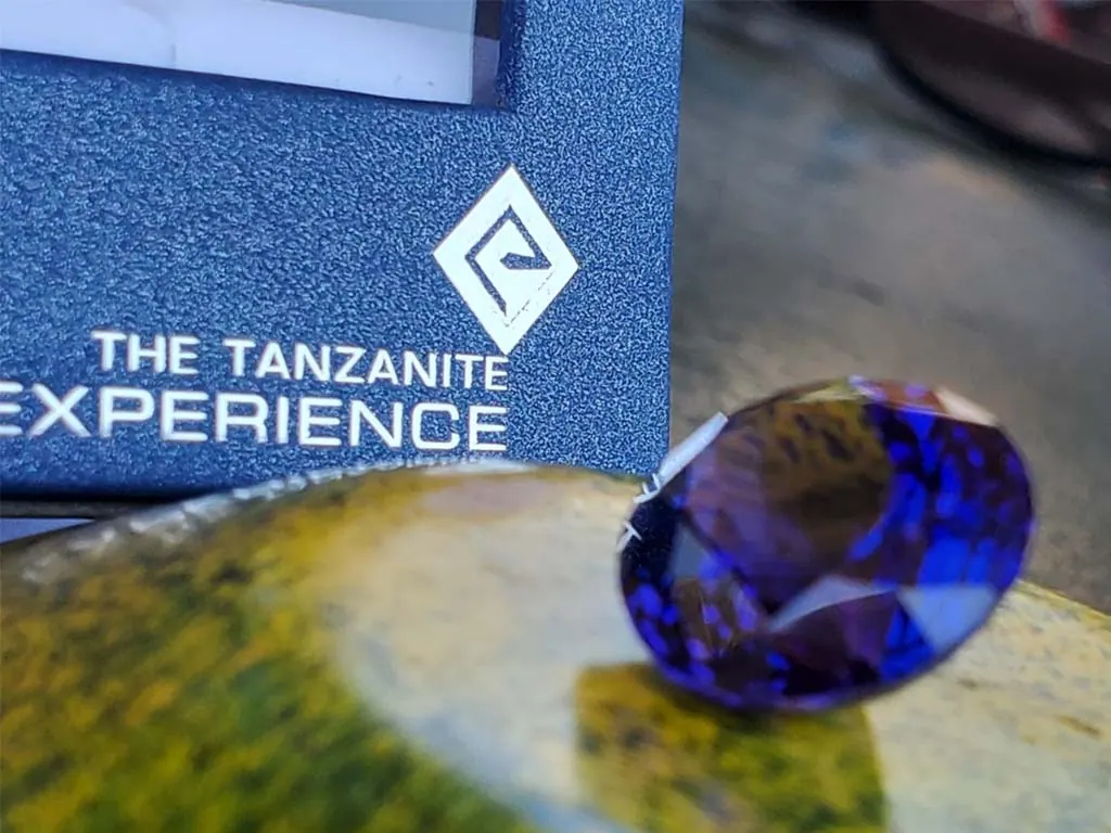A cut tanzanite gemstone on a polished surface beside The Tanzanite Experience box